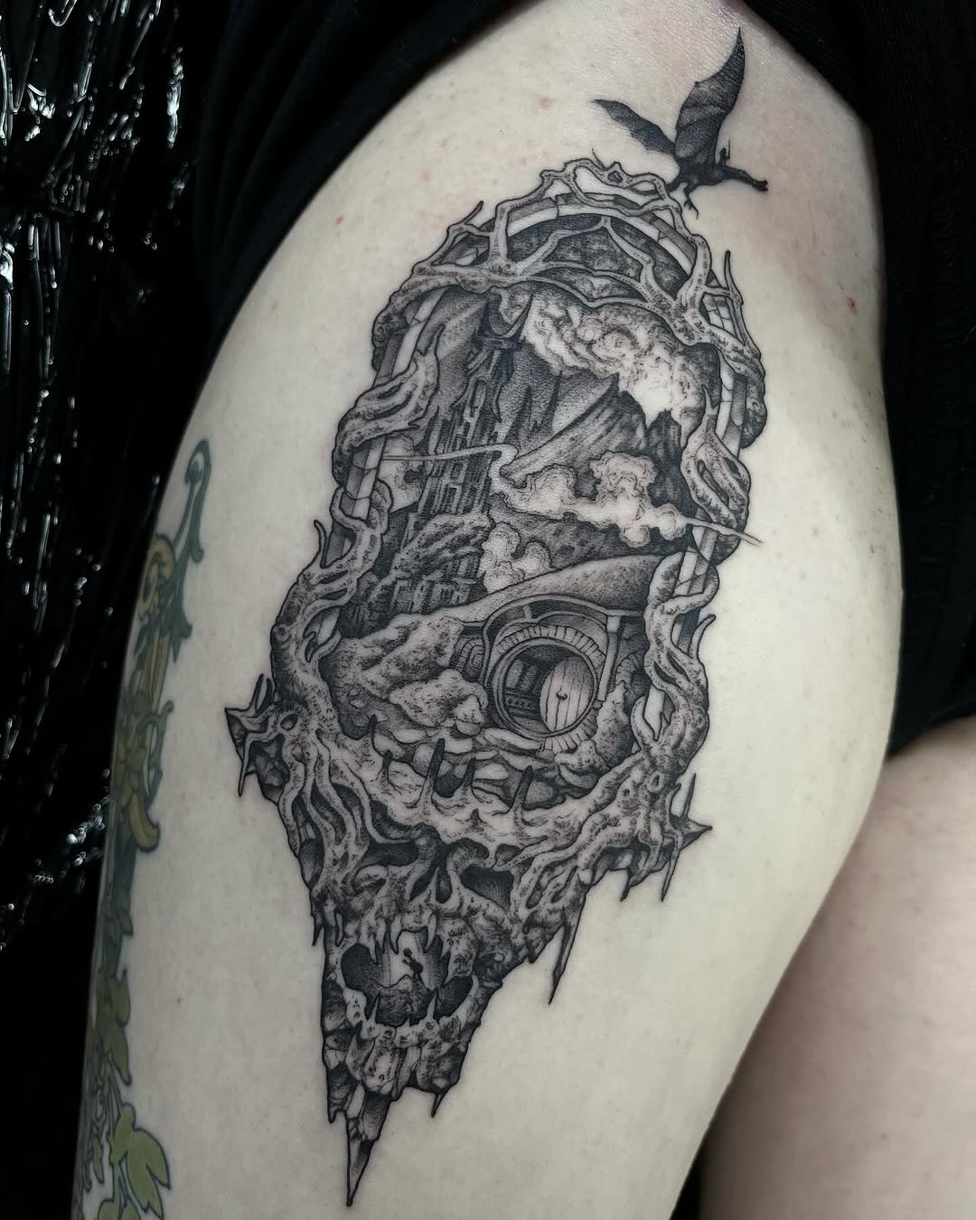 Detailed monochrome tattoo on upper arm featuring an ornate skull with organic branching elements, a bird at top, and an eye detail within the design.