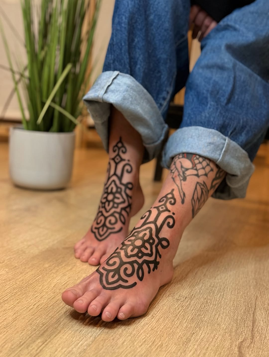 Ornate black floral and geometric foot tattoos featuring mandala-like patterns on both feet, displayed on a wooden floor.