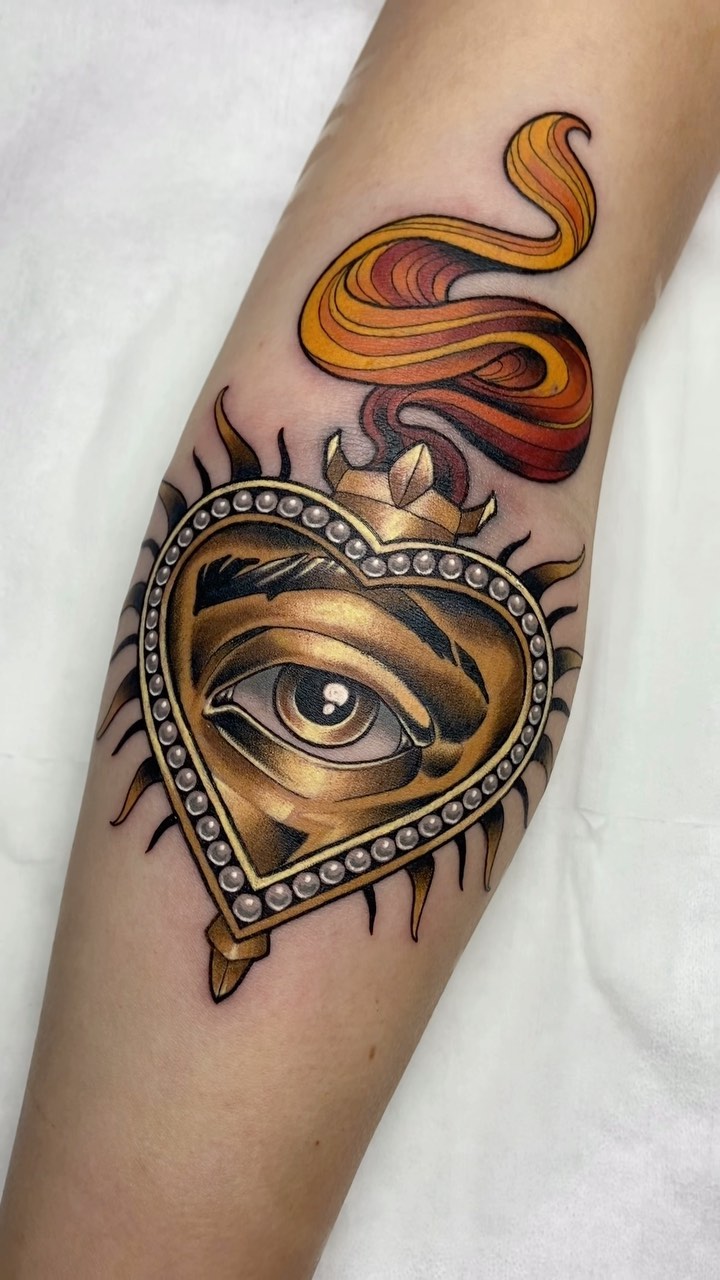 Golden heart with an eye and decorative pearls on the forearm, with flowing orange and red ribbons above in warm tones.