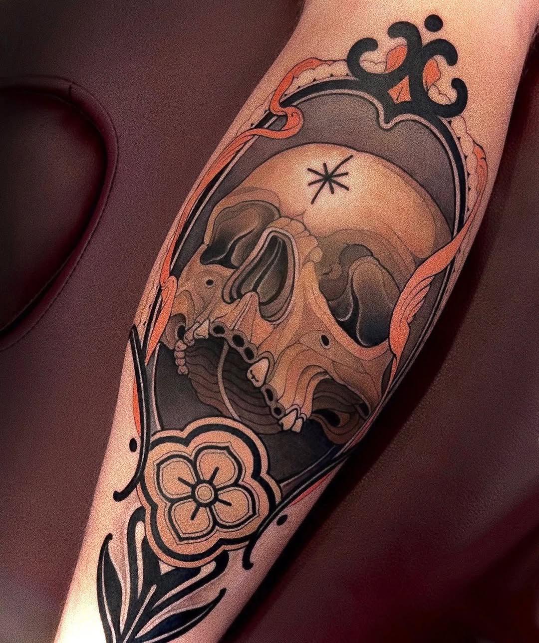 Skull tattoo on forearm with ornamental flourishes, flowers, and a crescent moon design in black, orange, and tan colors.