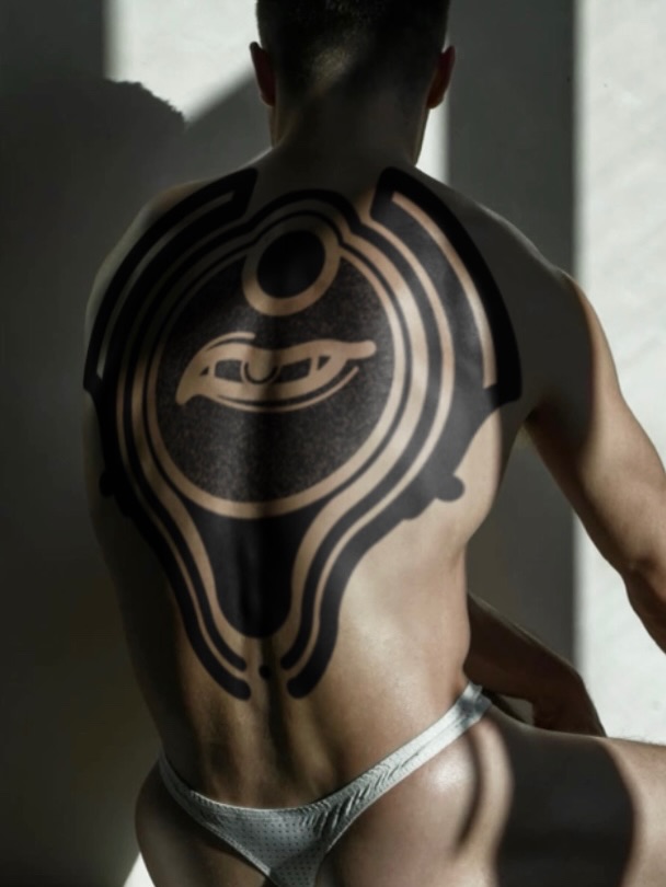 Shirtless man photographed from behind displaying a large geometric tattoo featuring concentric circles and curved lines in black and tan ink across his upper back.