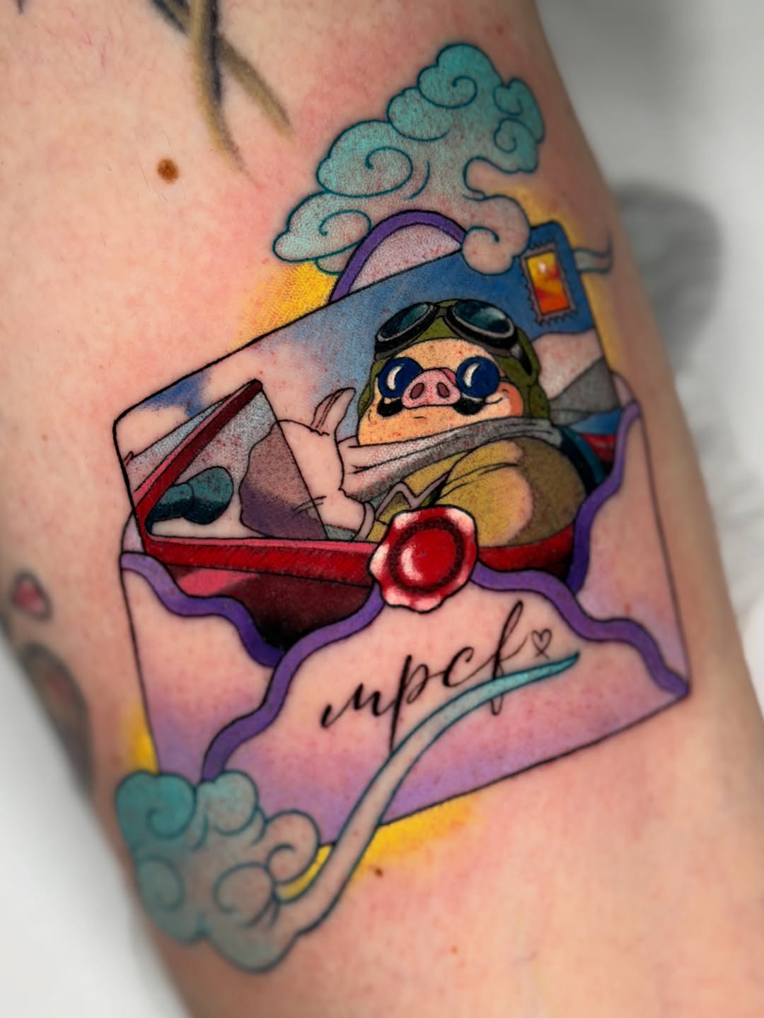 Colorful tattoo on an arm featuring a character in aviator gear inside a red airplane with purple clouds, yellow accents, and cursive text below.