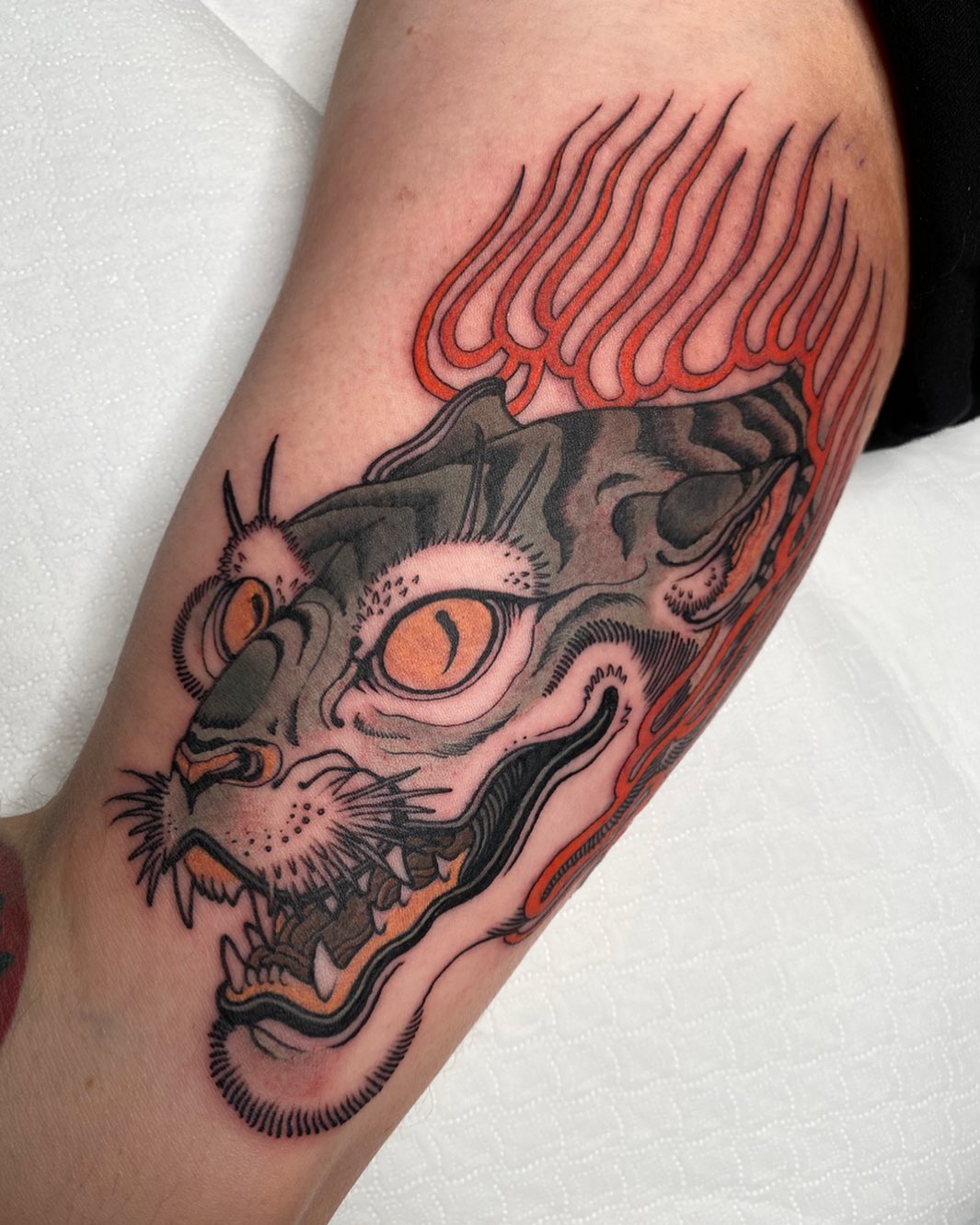 Colorful cat demon mask tattoo on upper arm featuring orange eyes, red and black flames, and intricate detailing in orange, black, and gray tones.