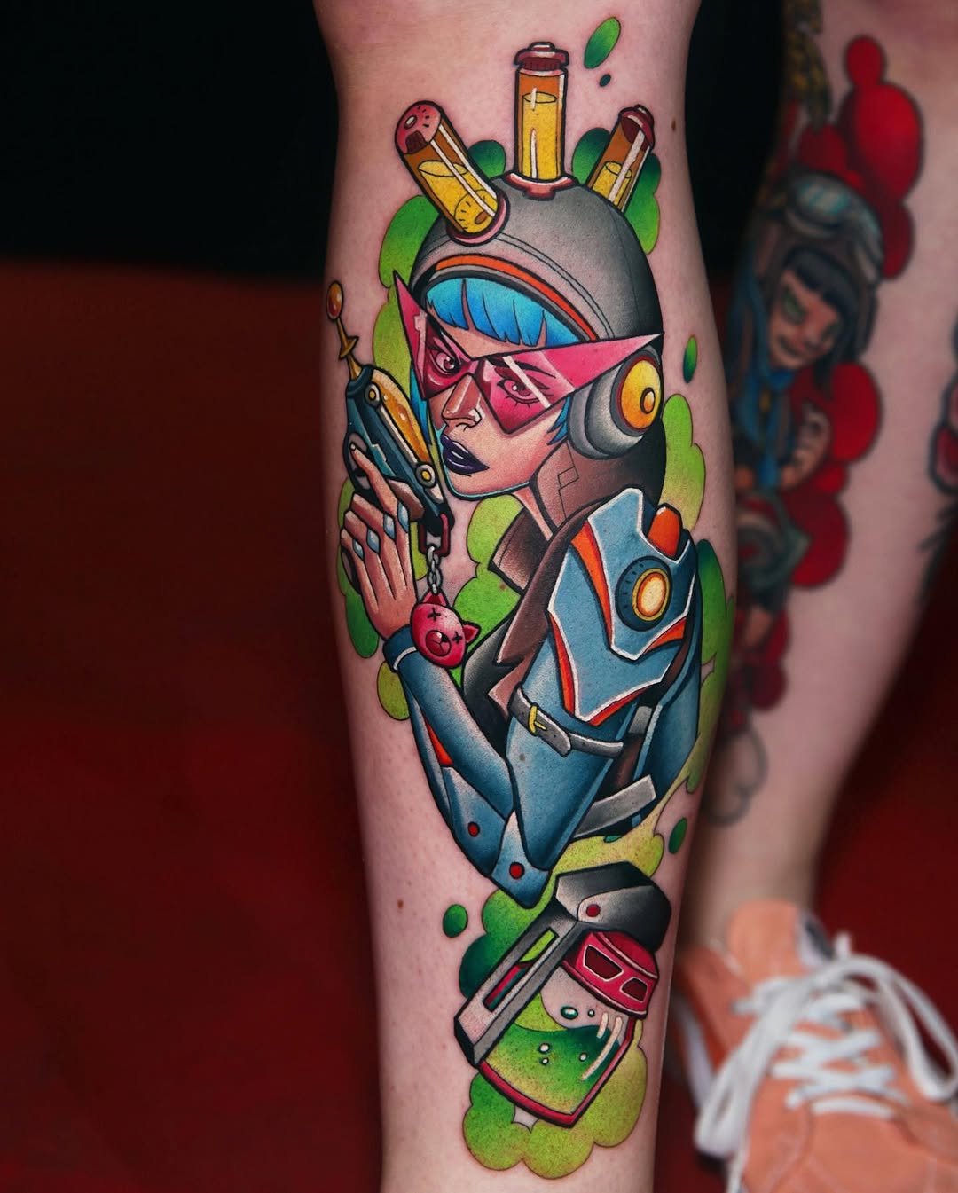 Colorful leg tattoo featuring a woman with pink mask and blue armor holding a gun, surrounded by bottles and green foliage.