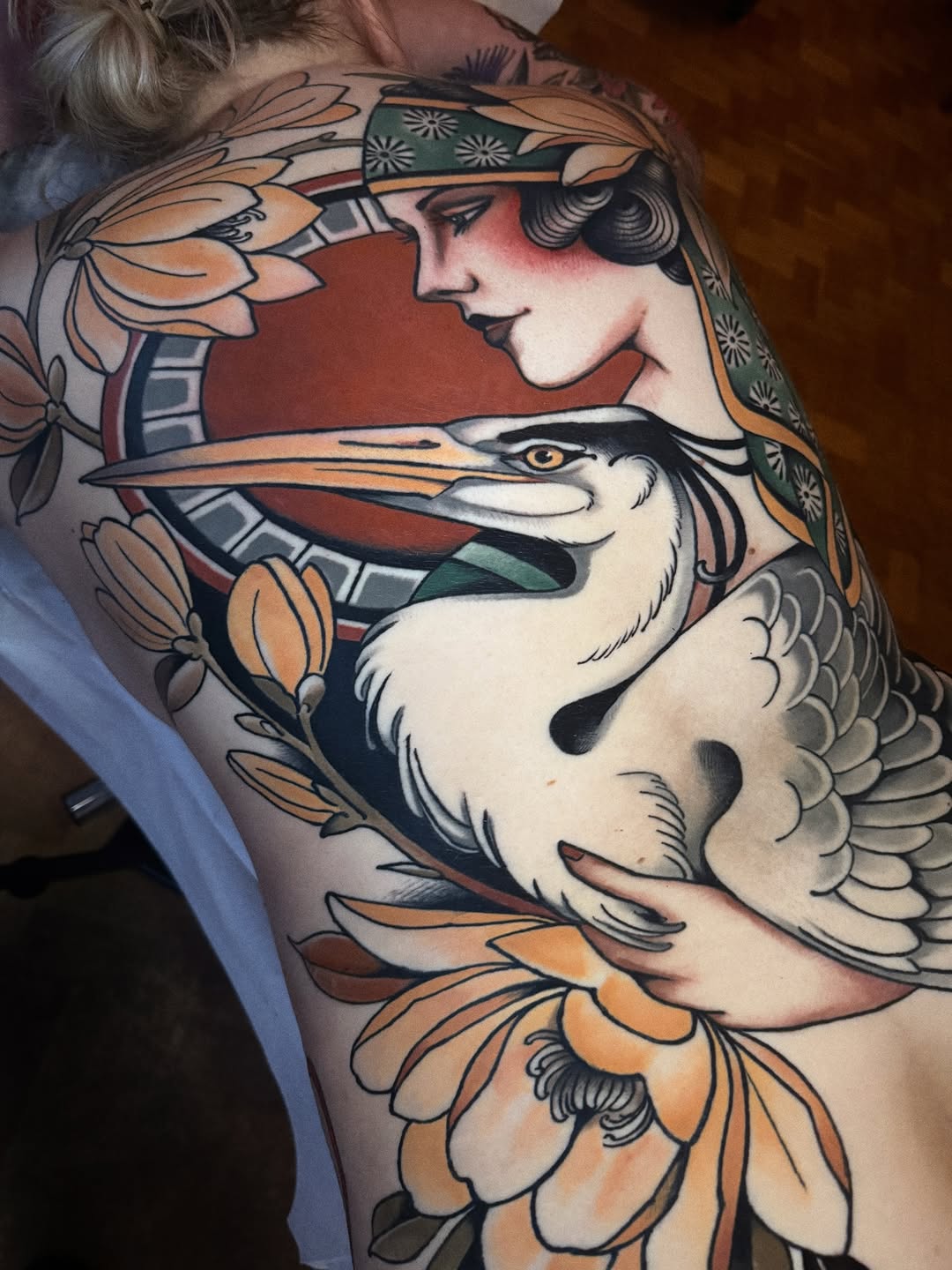 Colorful shoulder tattoo featuring a woman's profile with a headband alongside a crane, surrounded by orange flowers and decorative elements.
