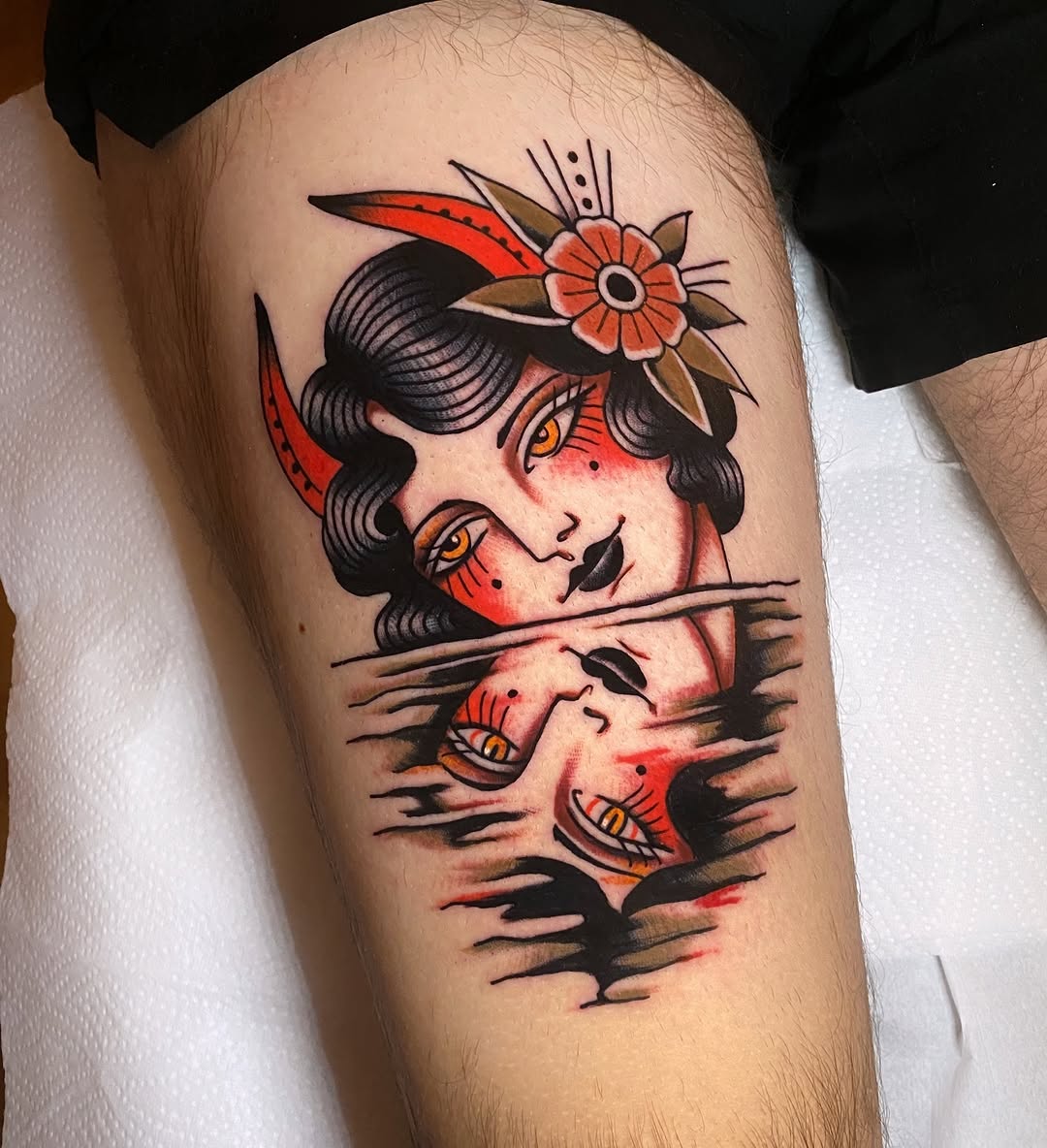 Colorful tattoo on upper arm featuring a red-faced woman with black hair, golden eyes, and flowers, with scratched texture effects below her face.