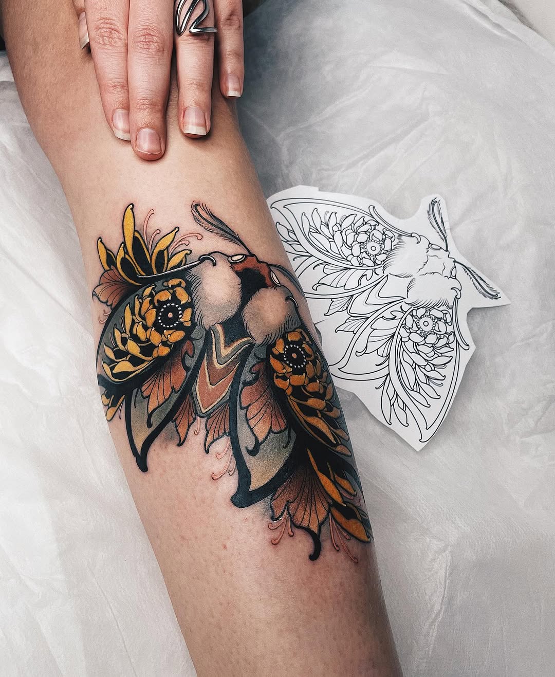 Colorful moth tattoo on forearm featuring orange, yellow, and black wings with floral details, displayed next to a line-drawn reference design.
