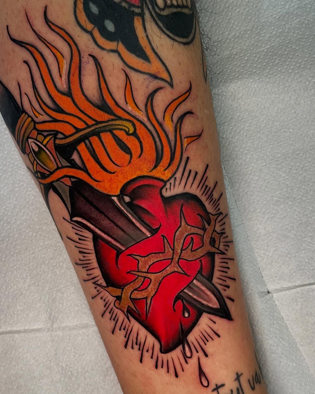Sacred heart tattoo on forearm featuring a red heart with orange flames, thorns, and radiating lines in vibrant color work.