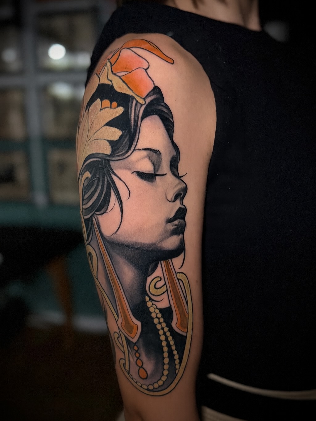 Profile portrait of a woman with closed eyes and ornate headdress featuring orange and black accents, displayed on upper arm with decorative elements including beaded necklace details.