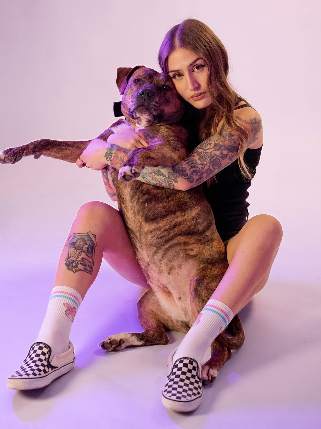 Woman with full arm tattoo sleeve featuring various designs in black and color, posing with a brown dog against a purple-lit background.