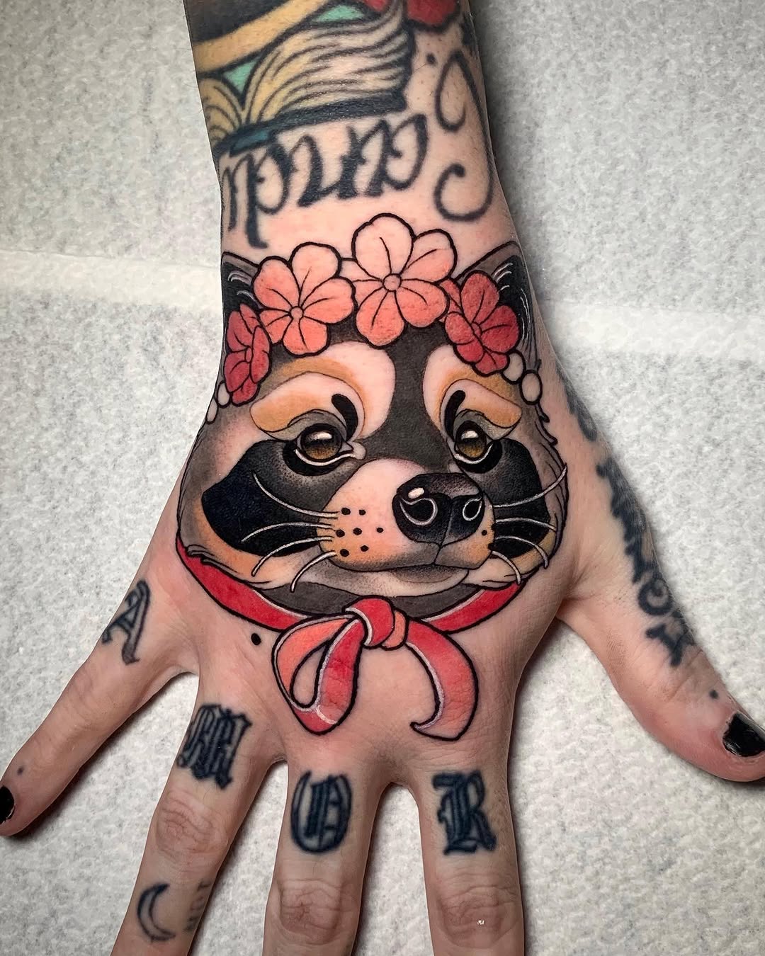 Colorful cat face with flower crown and red bow tattoo on hand, featuring black outlines, pink, red, and orange details.