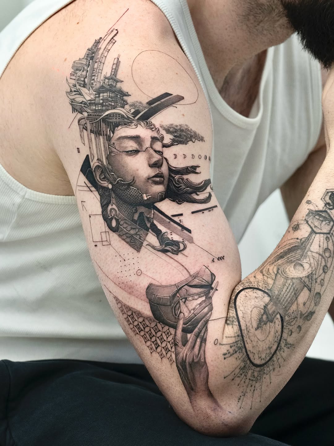 Monochromatic shoulder tattoo featuring a profile portrait with architectural and mechanical elements emerging from the head, geometric patterns, and decorative line work.