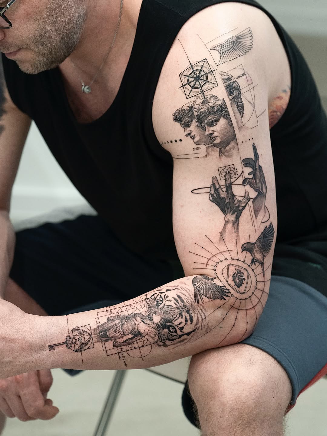Full arm tattoo sleeve featuring classical busts, geometric patterns, birds, a tiger, hands, and celestial symbols in black ink on pale skin.