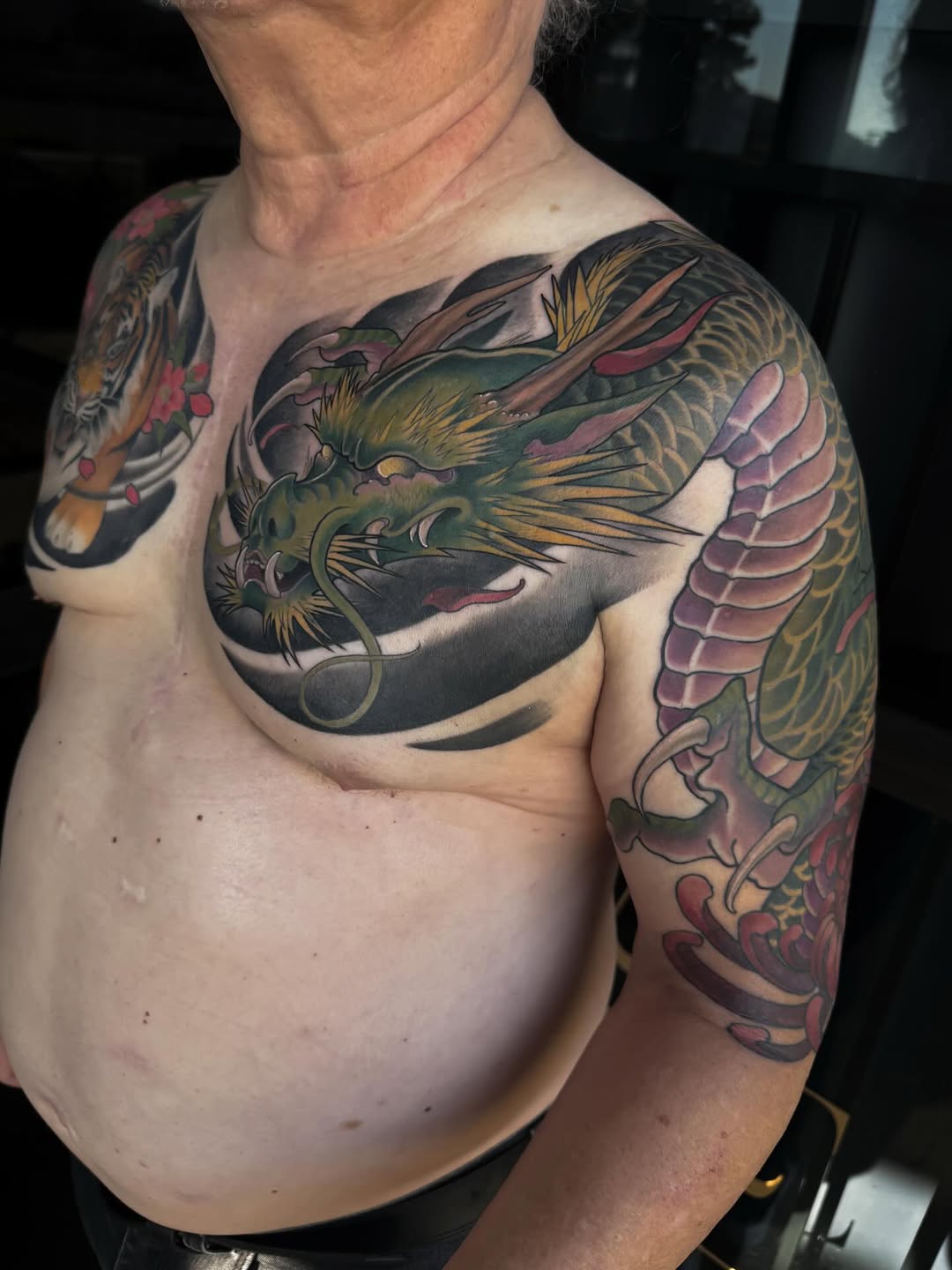 Colorful dragon tattoo wrapping across the chest and right shoulder with green, gold, pink, and purple scales and details.