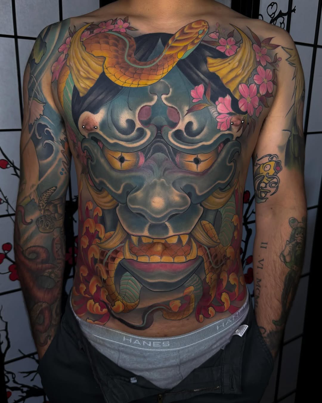 Large colorful chest and back tattoo featuring a horned demon mask with golden serpents, pink flowers, and ornate details in yellow, blue, and red tones.