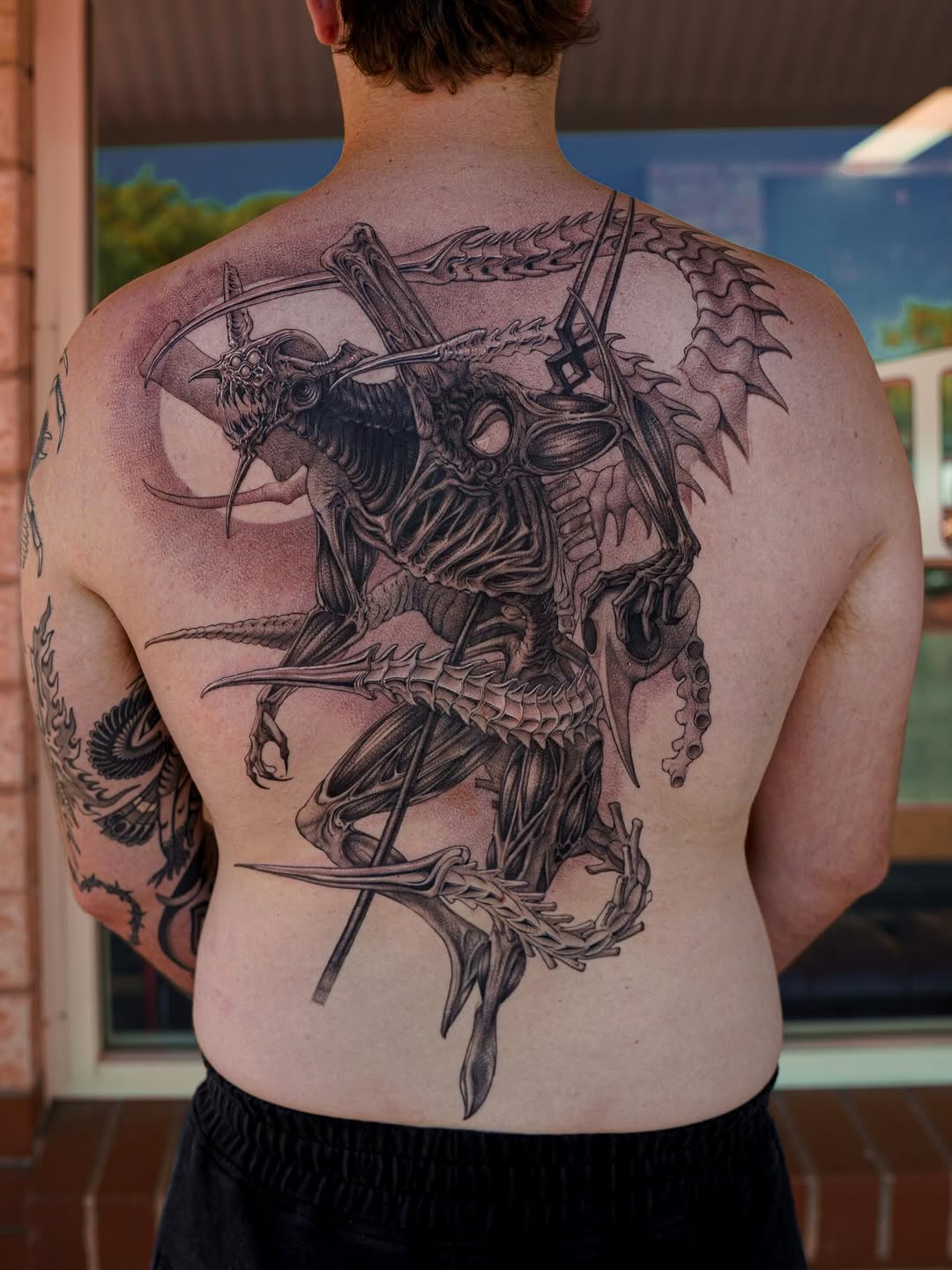 Back tattoo featuring a skeletal winged demon archer with detailed linework and shading in black ink on pale skin.