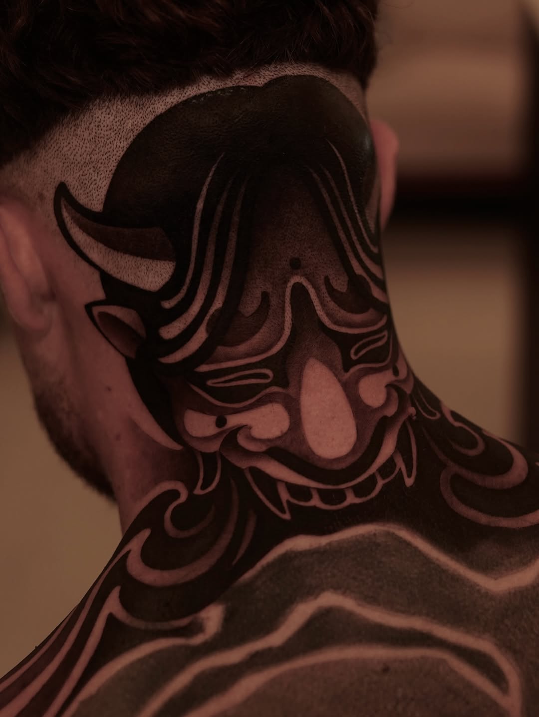 Ornamental demon or oni mask tattoo on the back of the neck featuring curved horns and decorative flourishes in black and red ink.