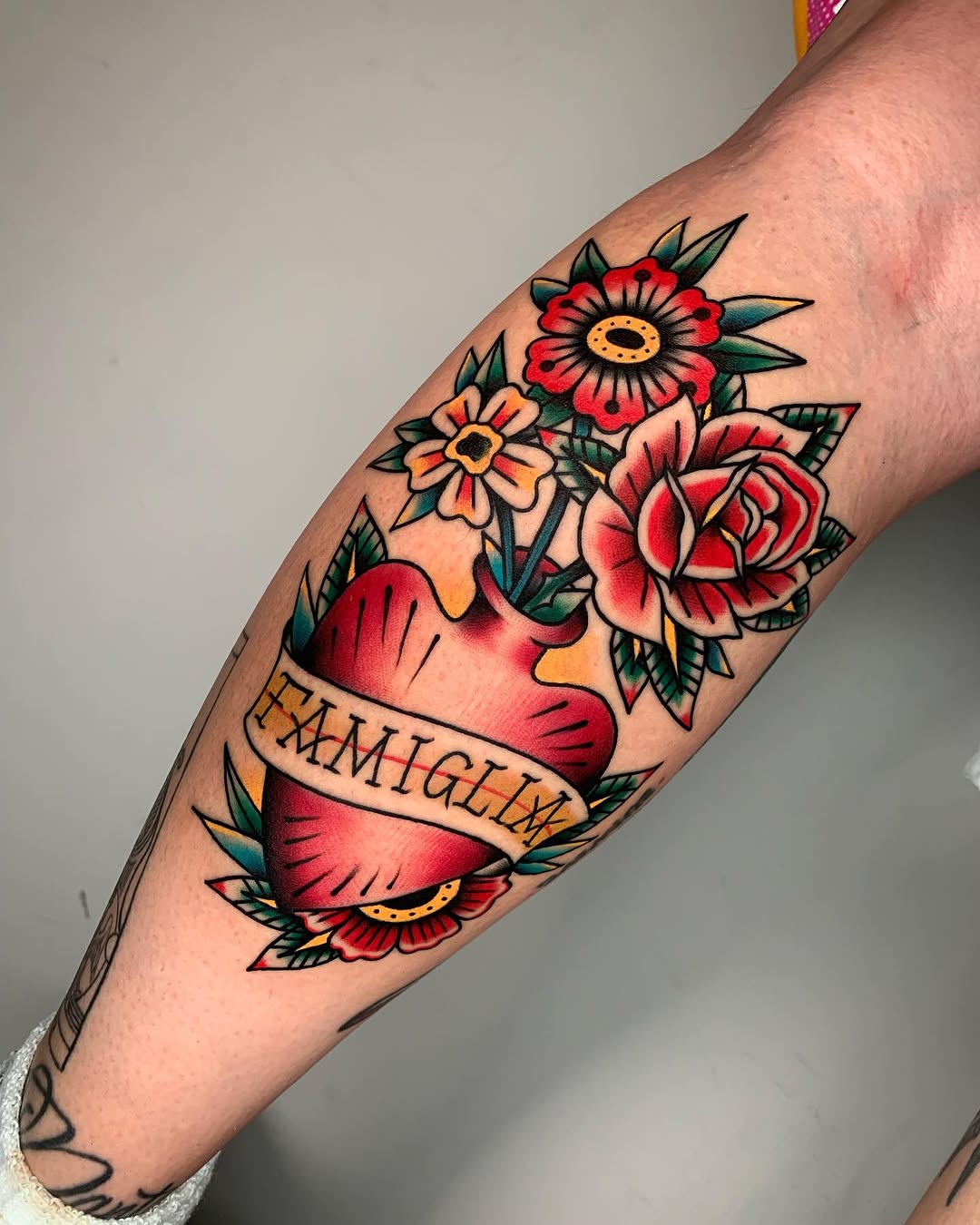 Red anatomical heart surrounded by colorful flowers including roses and daisies with green leaves and a banner reading "FAMILY" on the inner forearm.