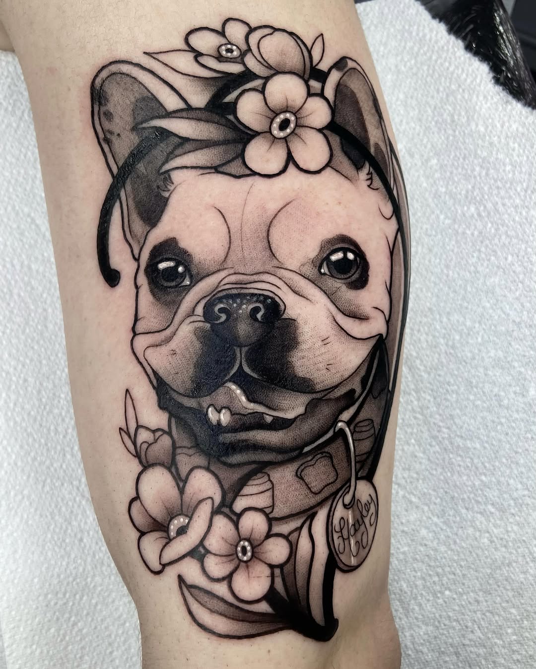 French Bulldog wearing a flower crown with decorative flowers and ribbons, tattooed in black and grey ink on the upper leg.