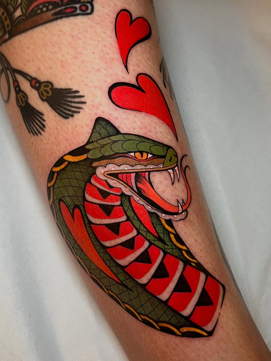 Colorful snake tattoo on forearm featuring a green head with orange and red striped body, red hearts above, and birds in upper background.