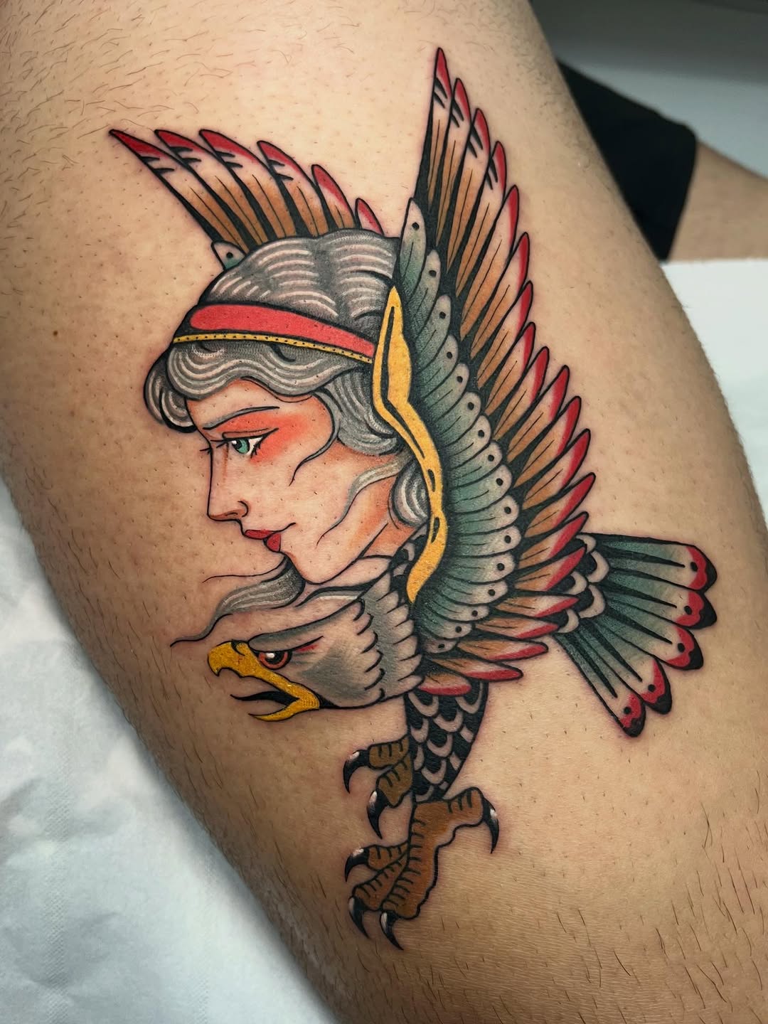 Colorful winged female figure with eagle head and body, wearing red headband, tattooed on upper arm with red, green, yellow, and flesh tones.