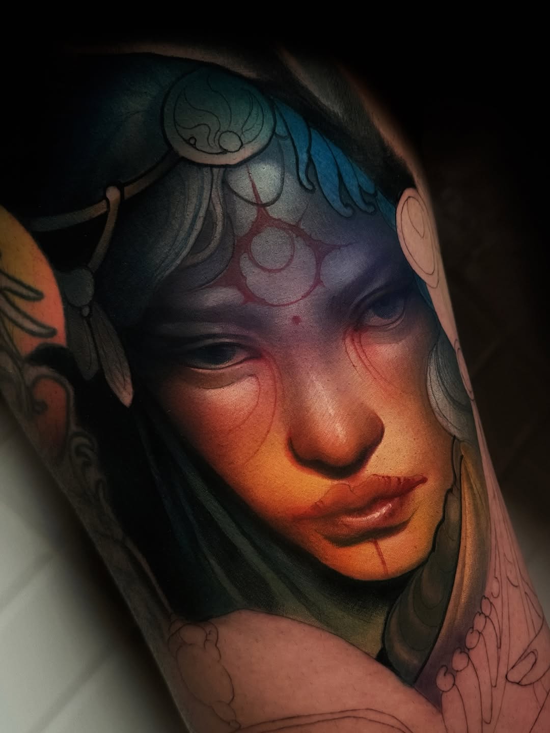 Colorful portrait tattoo on upper arm featuring a woman's face with blue and red facial markings, ornate headdress, and decorative line work.