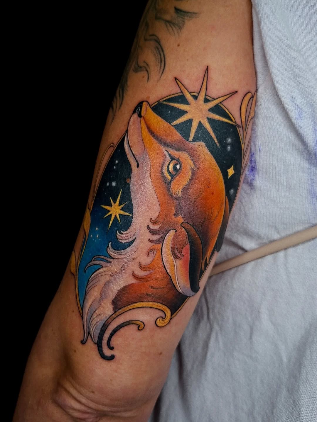 Colorful fox tattoo on upper arm featuring orange and red fur, dark starry background, gold stars, and decorative swirls in blue and black outlines.