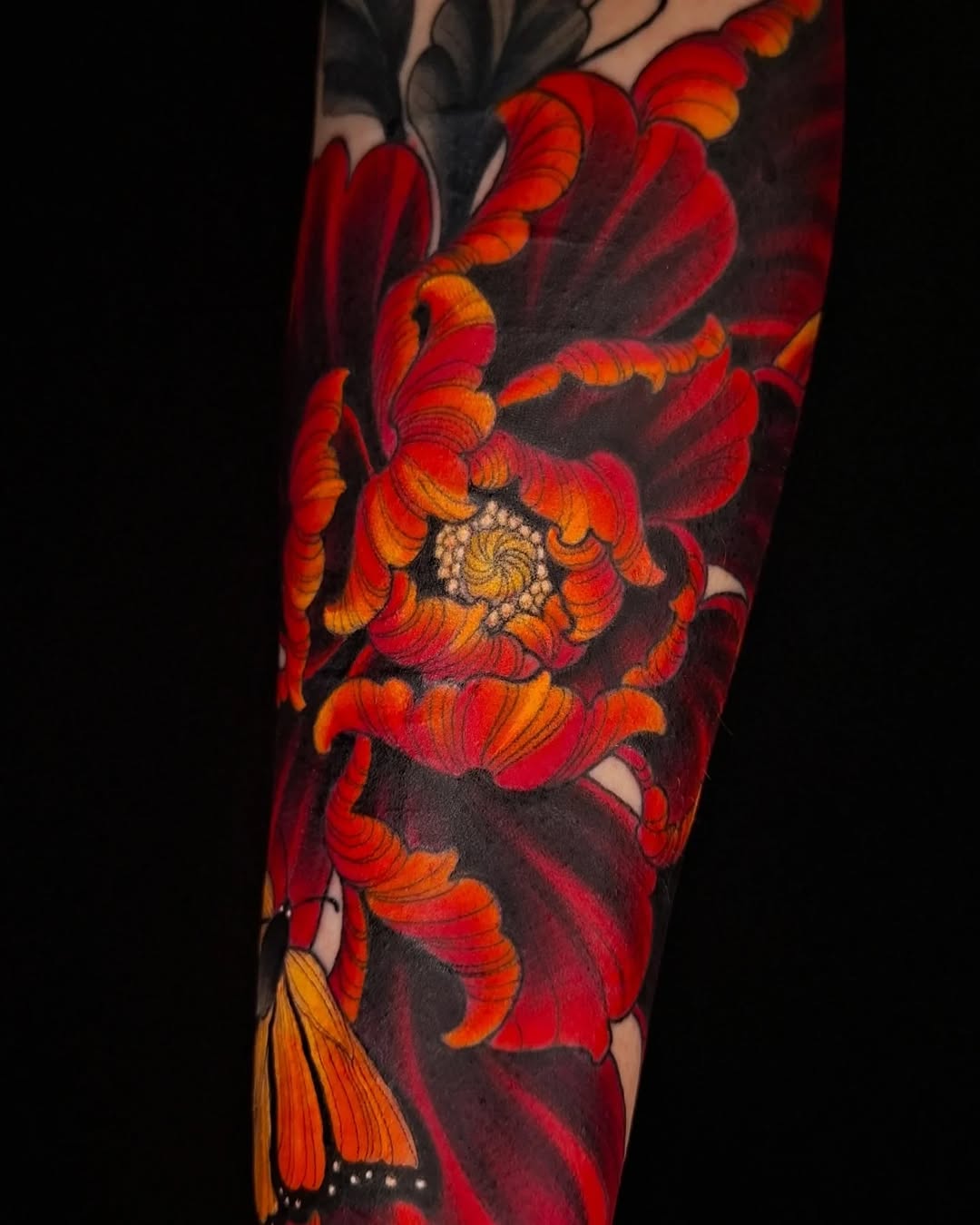 Vibrant red and orange flowers with a spiral center and decorative petals cascade down the forearm against a dark background.