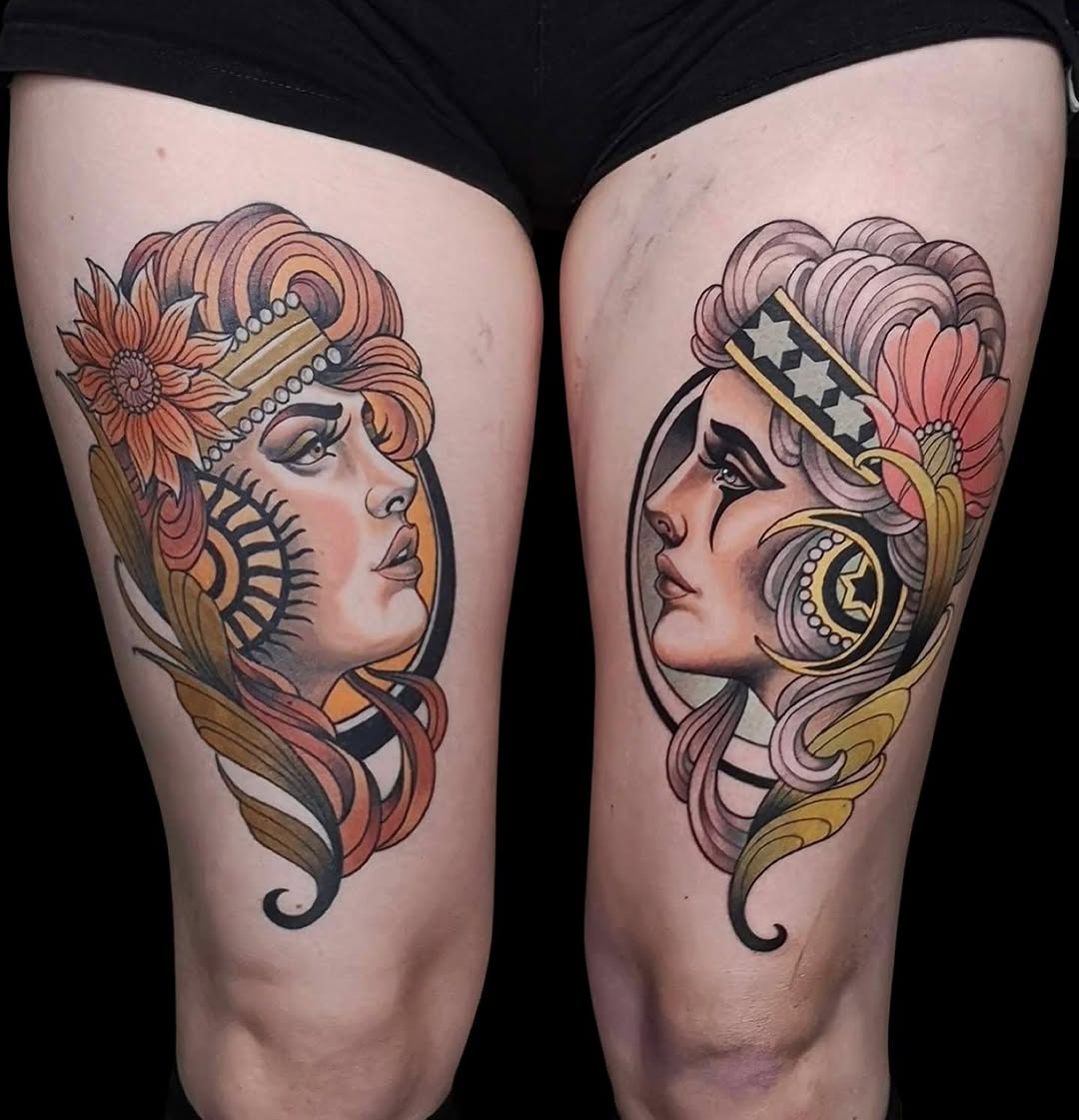 Two complementary thigh tattoos of stylized female heads with ornate headdresses, flowing hair, and decorative elements in coral, gold, and black ink.
