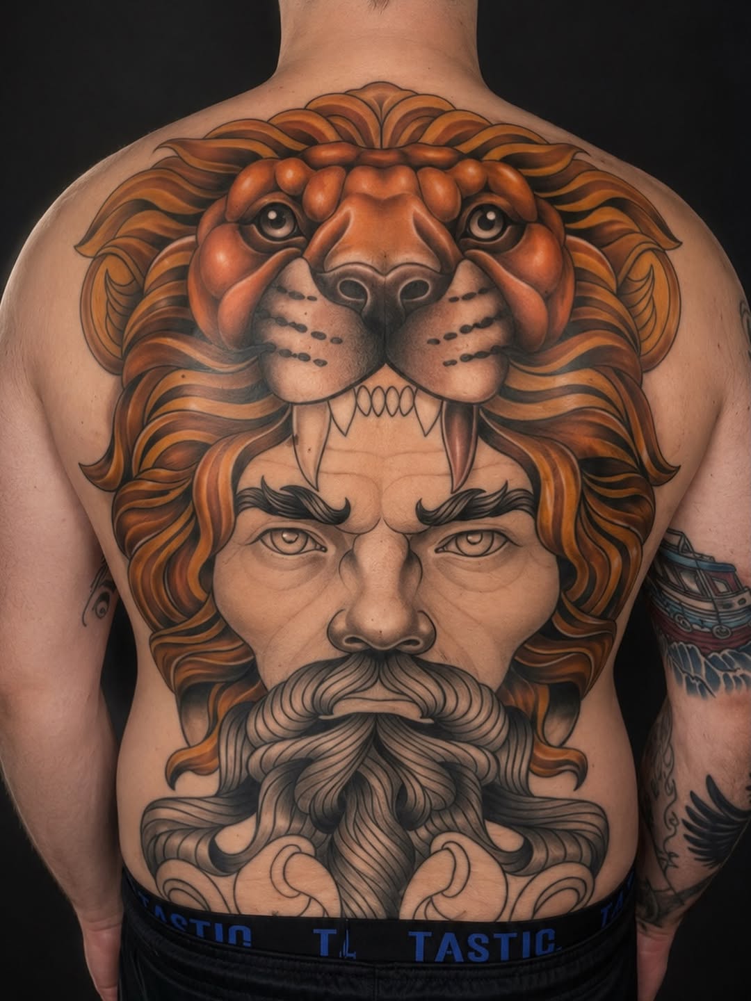 Large-scale back tattoo featuring a bearded man's face with a lion's head above it, rendered in orange and brown tones with black detailing.