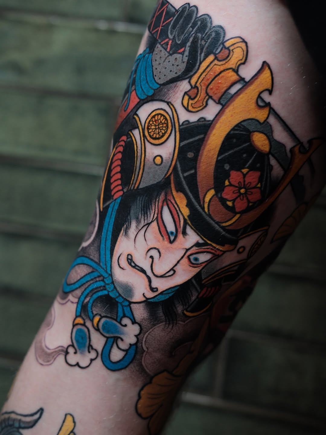 Colorful tattoo on an arm featuring a stylized face with blue and gold ornamental elements, holding a decorative object with red flowers and gold accents.