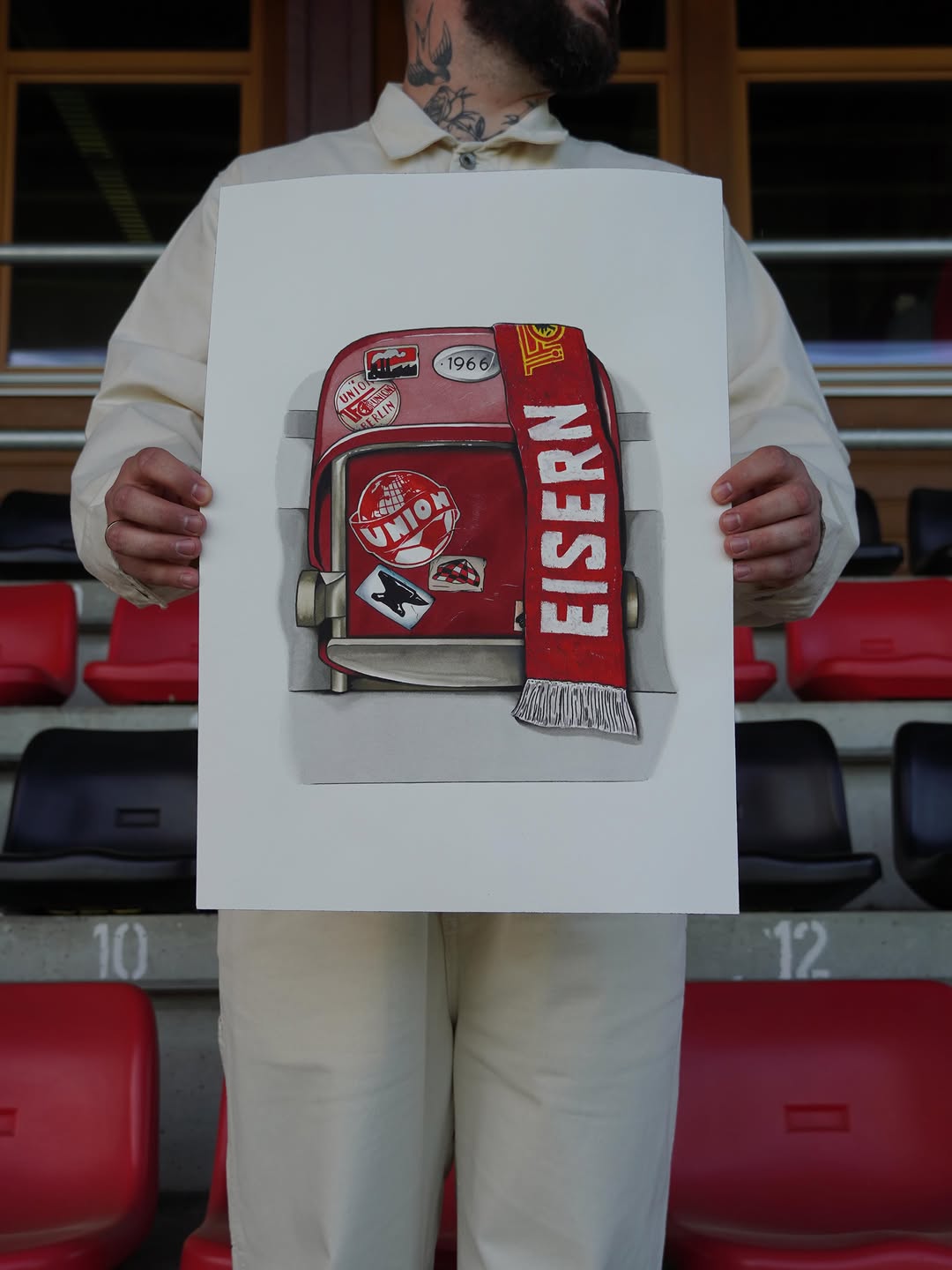 Person holding artwork displaying a red vintage suitcase with travel stickers and a red scarf labeled "CISERN" in a stadium setting.