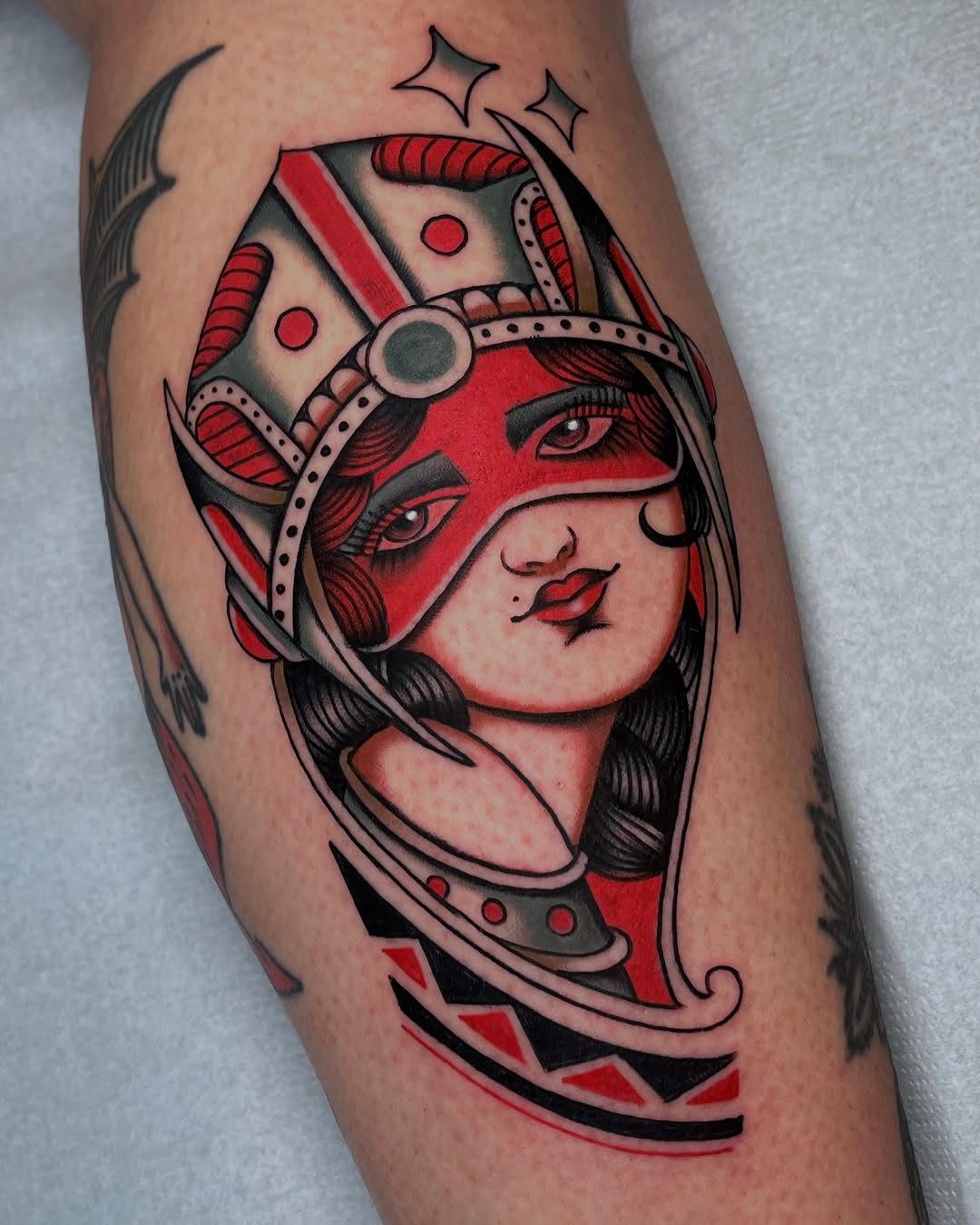 Woman's leg featuring a colorful tattoo of a red and black masked female figure with horns, decorative headpiece, and geometric patterns.