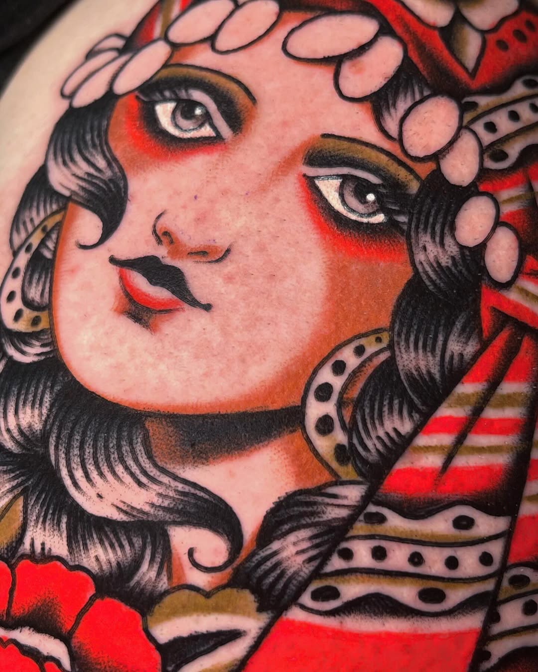 Colorful portrait tattoo of a woman's face with black wavy hair, red and orange shading, pink flowers in her hair, and decorative patterned elements on the shoulders.