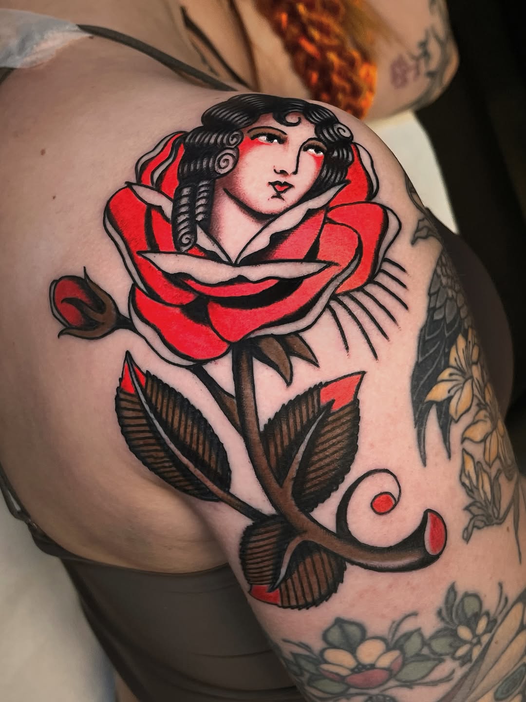 Woman's shoulder featuring a red rose with a female face emerging from the bloom, brown stem and leaves, surrounded by additional floral tattoos.