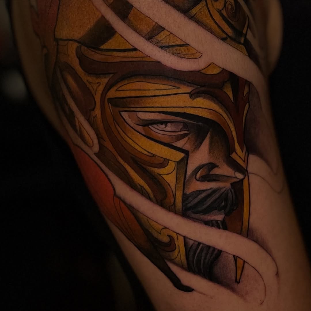 Detailed portrait tattoo of a bearded man wearing a golden helmet with dark shading, displayed on an arm with warm brown and gold tones.