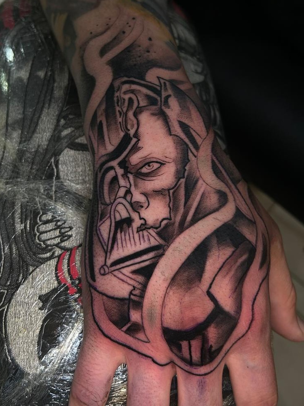 Hand tattoo depicting a demon or oni face with horns, shading in red and black ink, shown on a person's hand near fabric.