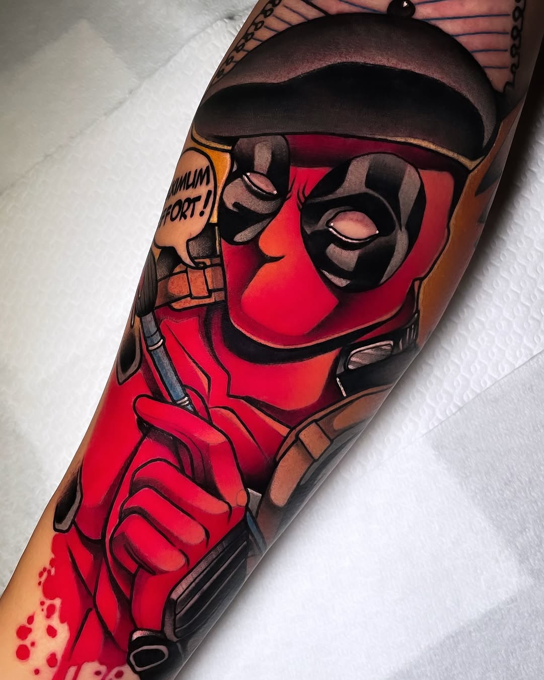 Deadpool character tattoo on forearm featuring red and black mask, brown tones, and yellow speech bubble with "MAXIMUM EFFORT" text.