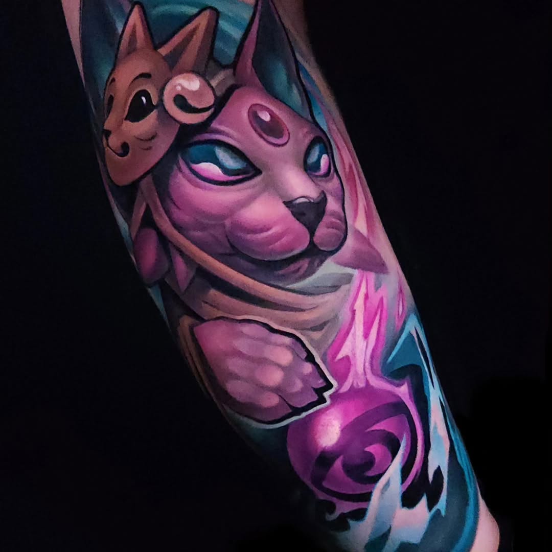 Vibrant pink and blue cat head tattoo with smaller cat mask design on upper arm, featuring detailed facial features and swirling decorative elements.