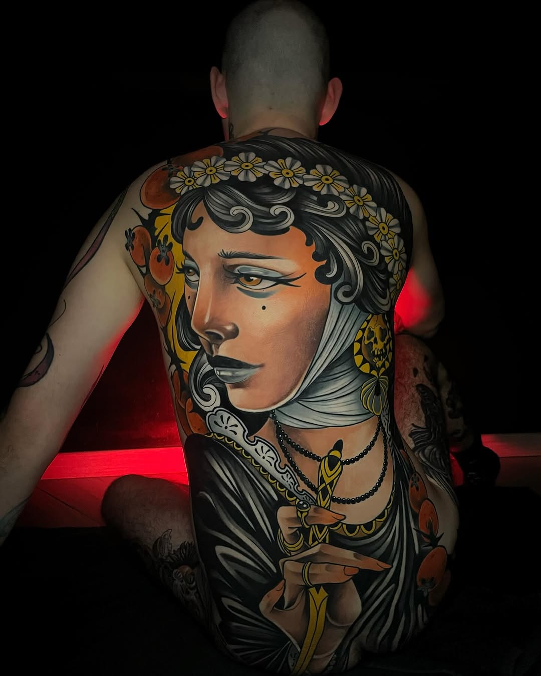 Large-scale portrait tattoo on back featuring a woman with flowing dark hair, daisy crown, and ornate jewelry in orange, gold, black, and cream tones.