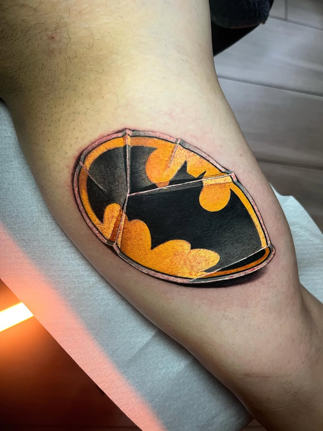 Batman logo shield tattoo on the upper arm featuring black and yellow colors with orange and red shading and outlined details.