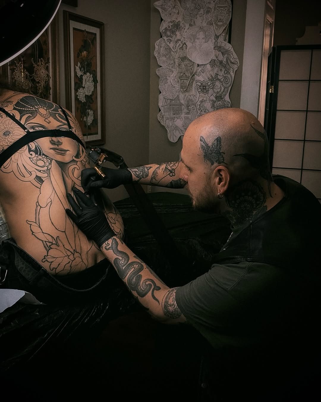 Tattoo artist applies ink to client's upper back and shoulder, featuring intricate botanical and portrait designs in black and grey tones.