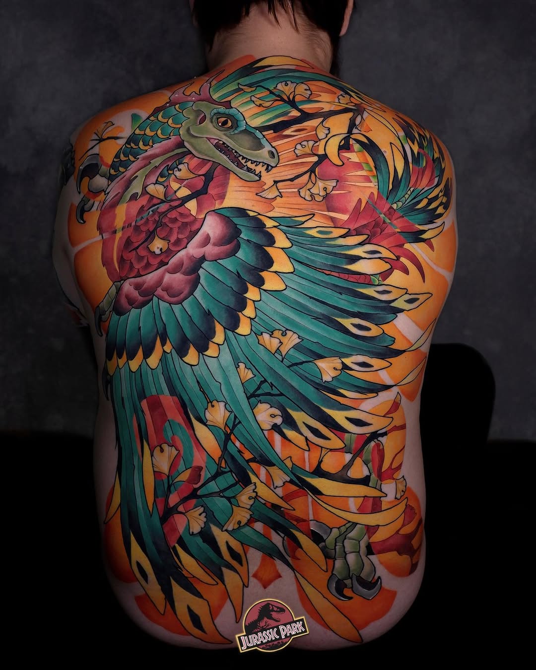 Colorful back tattoo featuring a winged dragon with orange, teal, and red hues, scales, and flowing feathers covering the upper back and torso.
