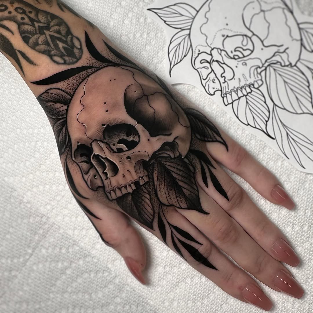 Skull with floral leaves and botanical details tattooed on the back of a hand, shown in black and gray ink alongside a line-drawn design sketch.