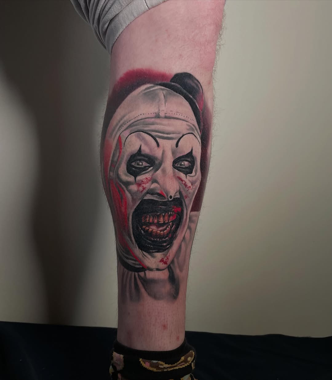 Horrified clown face tattoo on calf with black ink, red shading, and expressive eyes with dramatic mouth expression.