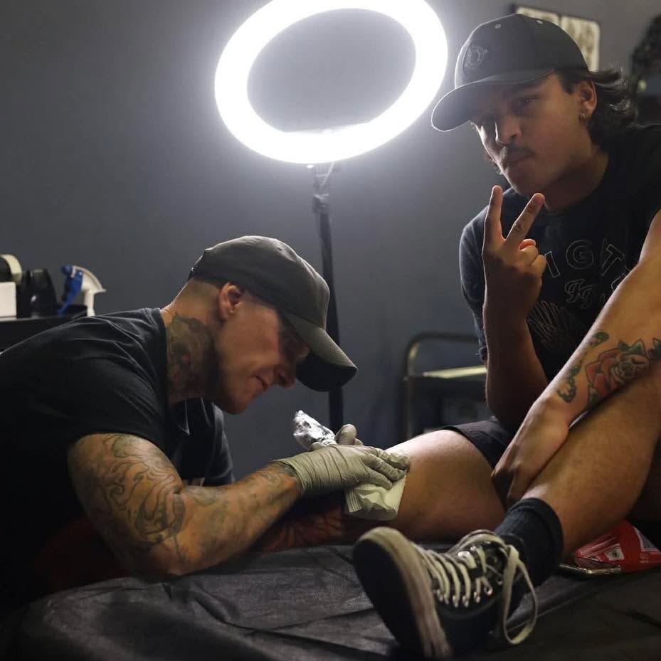 Tattoo artist with tattooed arms wearing a cap applies ink to a client's leg under a bright ring light in a studio setting.