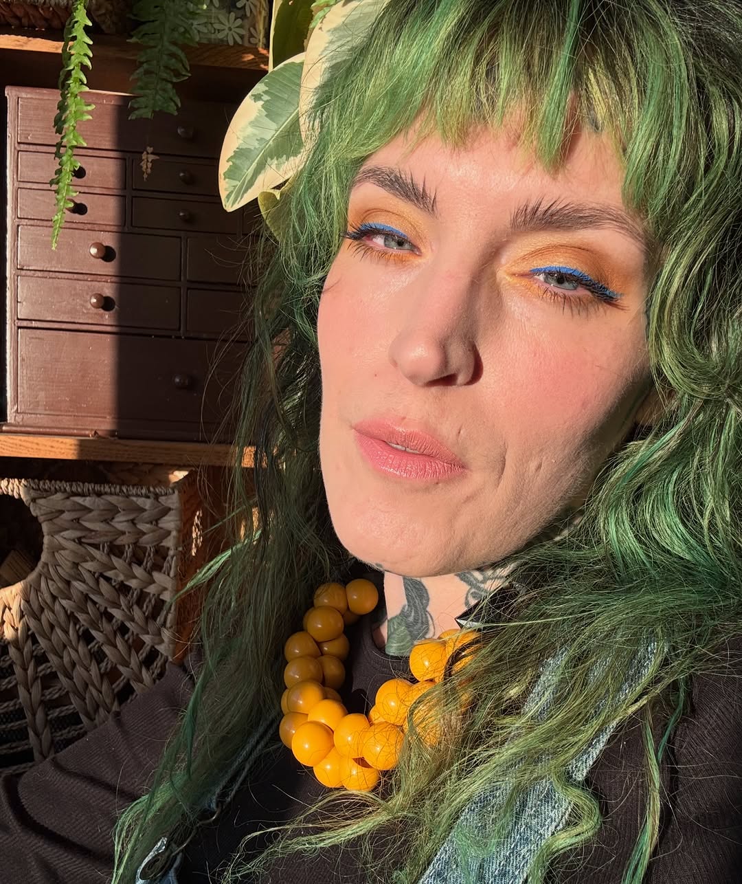 Person with green hair displaying a gray geometric tattoo on their neck and yellow-orange beaded necklace.