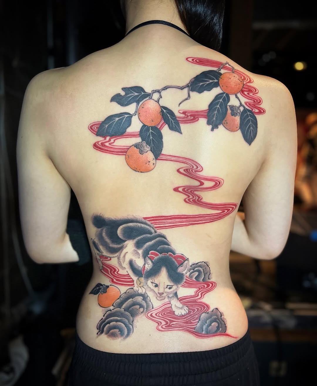 Back tattoo featuring orange fruits on branches with dark leaves connected by flowing red wavy lines to a cat figure with clouds below.