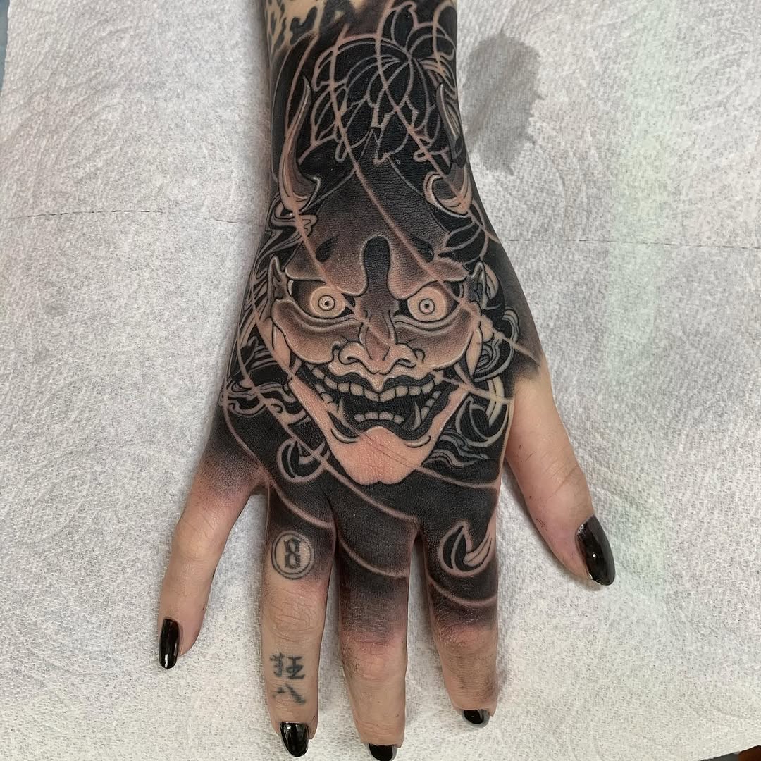Demon face tattoo with ornate floral details covering the hand and forearm in black and grey ink.