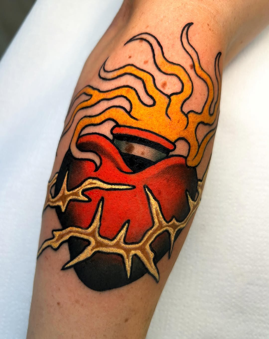Sacred heart tattoo on an arm featuring a red heart with flames in orange and yellow, surrounded by a gold thorned crown and black shadows.